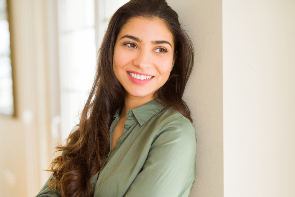 A woman with long hair leans comfortably against a wall, smiling softly and showcasing clear, healthy vision in a naturally lit indoor environment. - Eye Exams