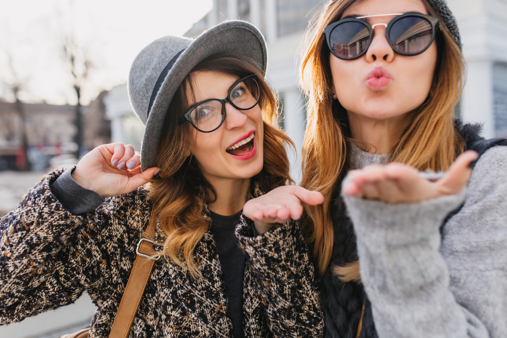 Two stylish young women smile and blow kisses at the camera, one wearing chic eyeglasses and a hat while the other sports bold dark sunglasses, capturing a fun and fashionable urban moment – Sunglasses.