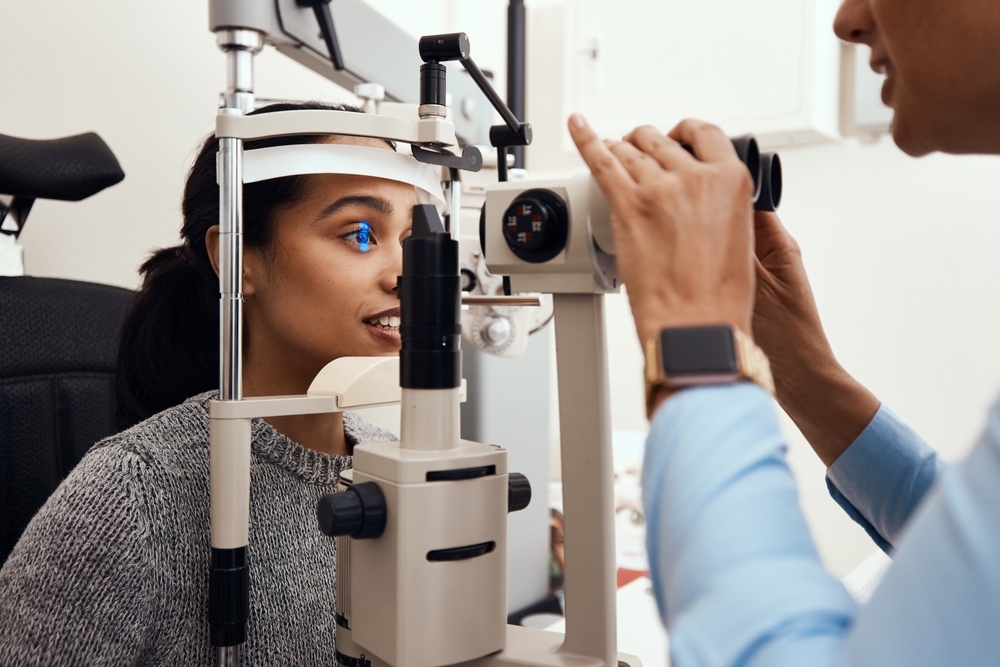 A woman sits at an exam machine with a blue diagnostic light reflecting in her eye while an eye care professional conducts a detailed evaluation. The setting highlights modern eye-care technology focused on precision and comfort – Neurolens.