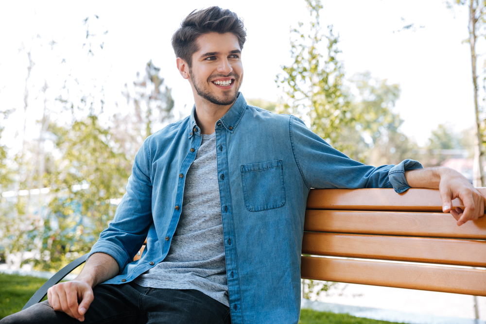 A cheerful young man sits comfortably on a park bench, enjoying the sunshine and natural surroundings. His bright, confident smile creates a warm and positive atmosphere, symbolizing clear vision and overall eye health – Cataract Treatment.