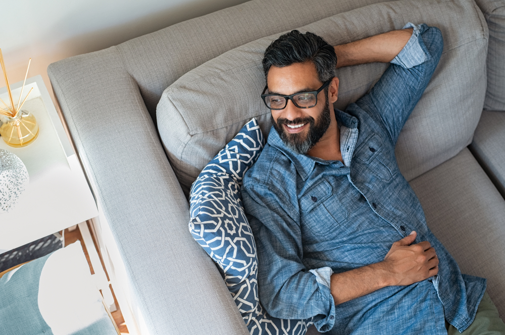 Relaxing Man with Modern Prescription Glasses – Eyeglasses A bearded man lounges comfortably on a sofa while wearing fashionable eyeglasses. His relaxed posture and confident smile showcase both comfort and clear vision – Eyeglasses.