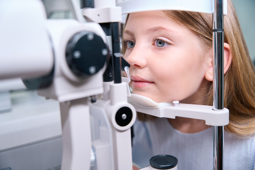 A young girl sits at a slit-lamp machine during an eye examination, smiling slightly as she looks forward while the optometrist evaluates her eye health. The clinical setting emphasizes early vision care – Contact Lenses.