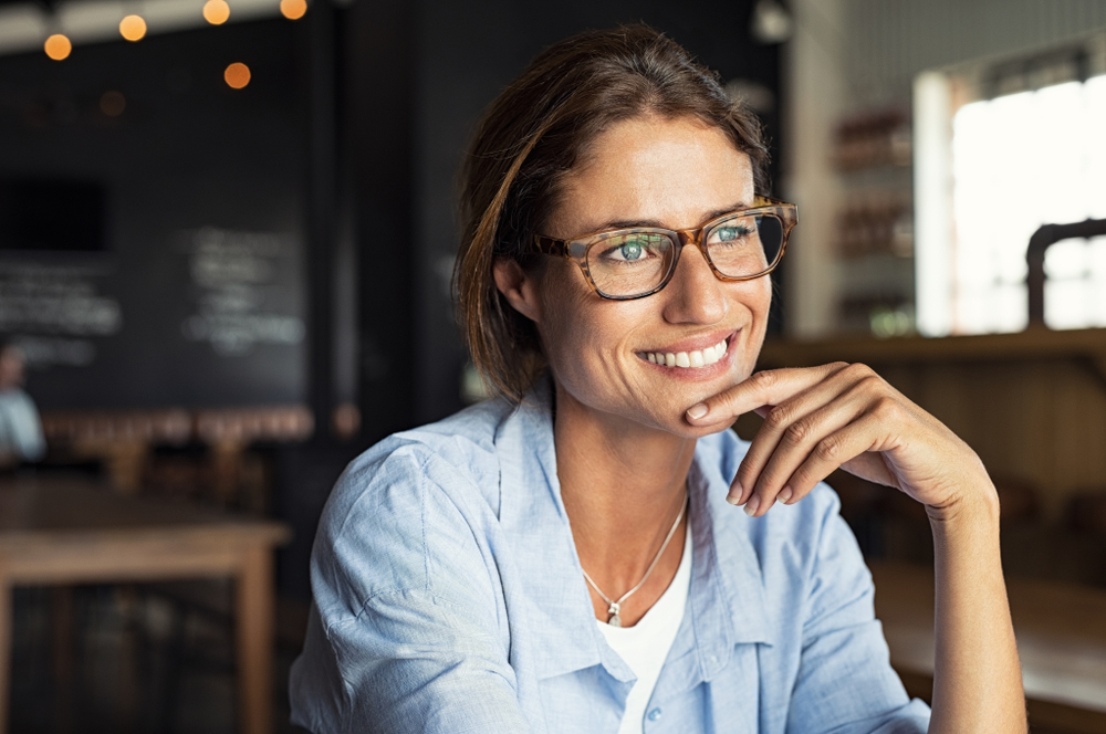 A confident woman wearing fashionable eyeglasses smiles warmly while seated in a cozy café. Soft lighting and a relaxed atmosphere emphasize clarity, comfort, and modern visual wellness – Neurolens.