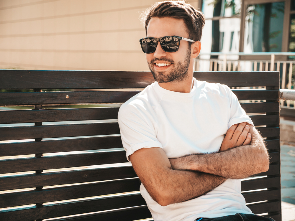 A man lounges comfortably on a wooden bench with his arms crossed, confidently wearing sleek black sunglasses and a white shirt while enjoying a warm, sunny day – Sunglasses.