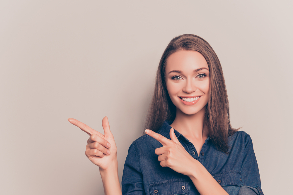 A cheerful woman with clear, bright eyes points to the side with both hands, standing against a neutral background. Her confident smile and eye contact create a welcoming, promotional feel – Contact Lenses.