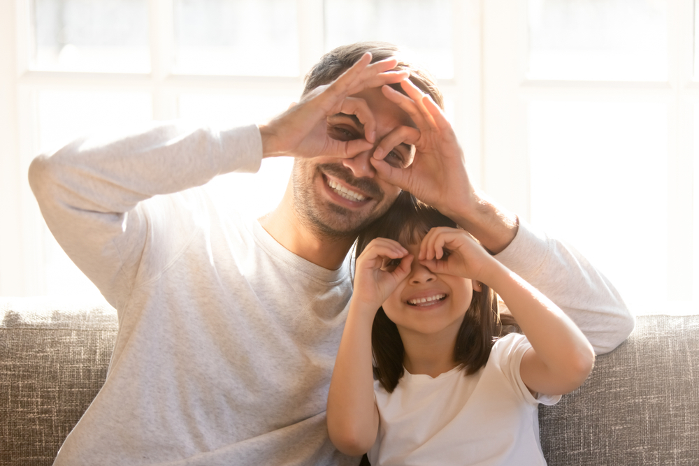 A joyful father and daughter sit together on a couch, forming playful “eye glasses” with their hands as they smile toward the camera. The bright natural light creates a warm, happy atmosphere, capturing the fun and connection between them – Neurolens.