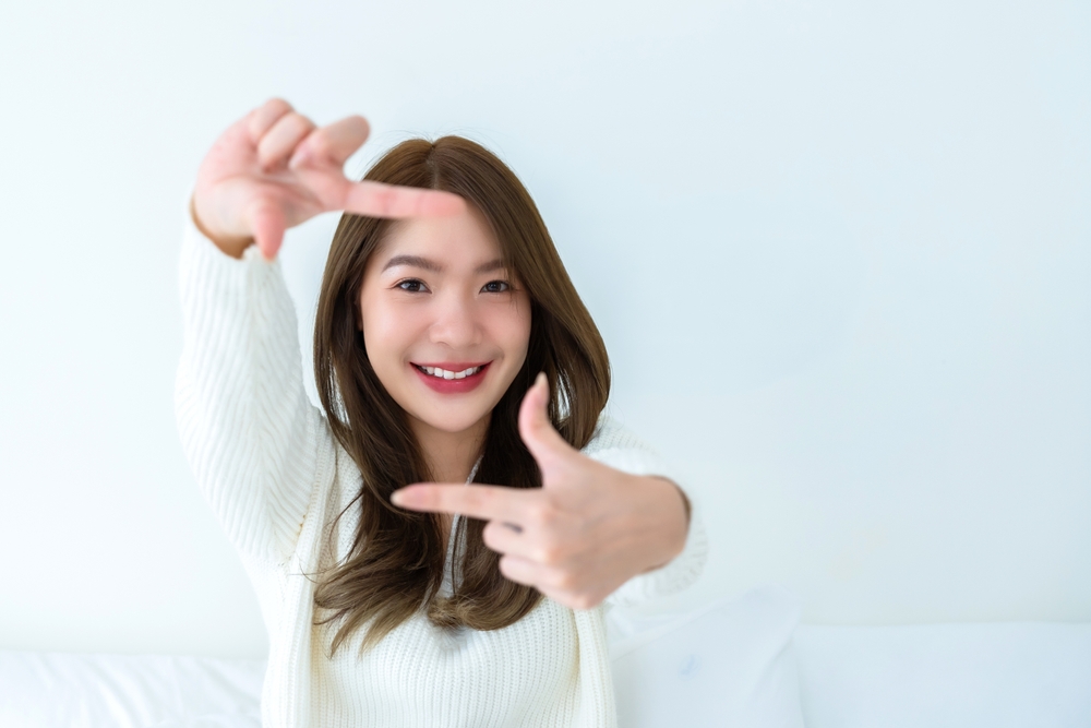 A cheerful young woman uses her hands to form a frame around her face, emphasizing her bright, healthy eyes while smiling playfully in a bright, minimalist setting. - Eye Exams