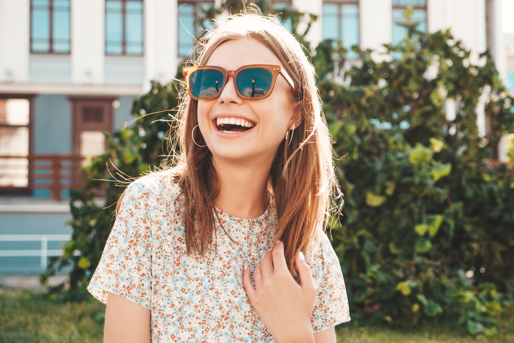 A cheerful young woman wearing trendy brown-framed sunglasses laughs brightly while outdoors, with sunlight illuminating her face and hair, capturing a carefree, sunny-day moment – Sunglasses.