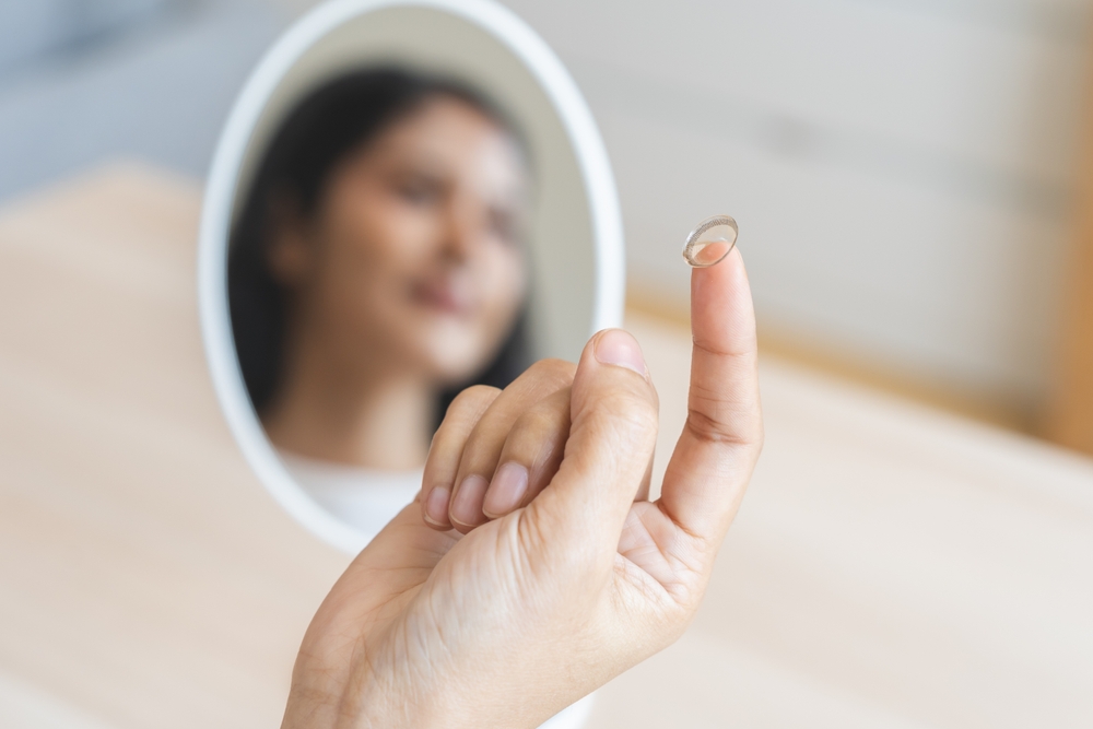 A close-up of a woman holding a clear contact lens on her fingertip while looking into a mirror, preparing for insertion. The soft, natural lighting highlights the careful placement and daily contact lens routine – Contact Lenses.