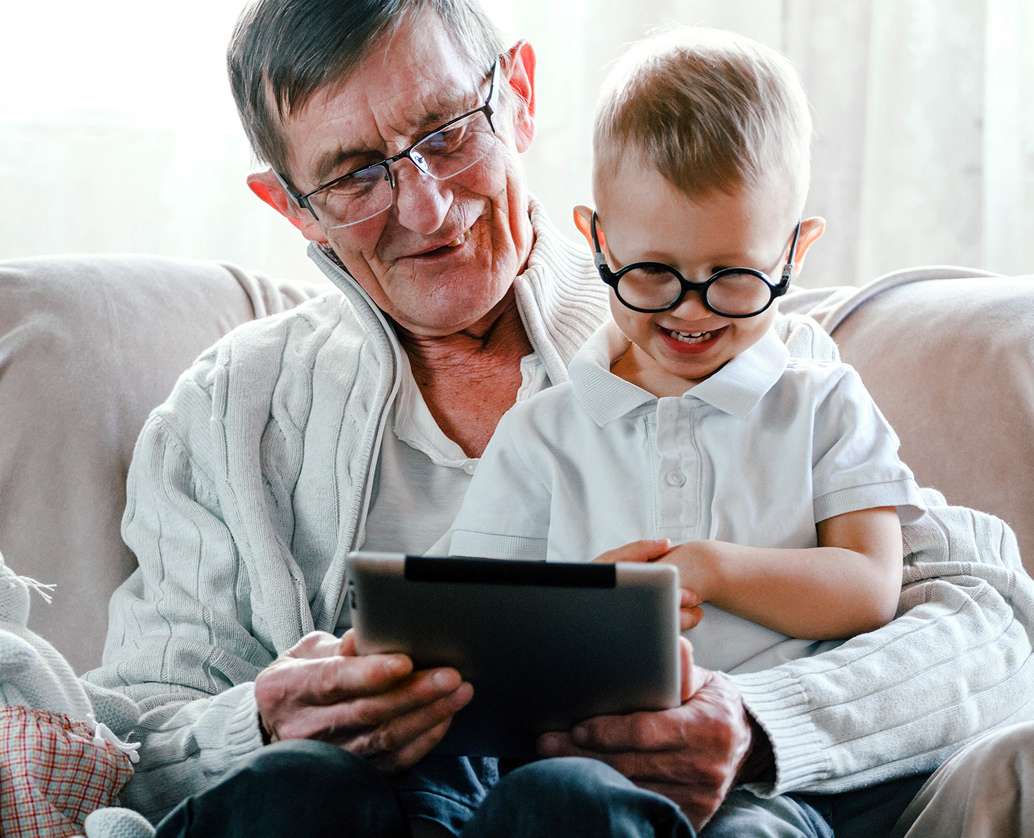 An elderly man and a young boy, both wearing glasses, smile as they look at a tablet together while sitting on a couch - Eye Doctor Rolla MO | Invision Family Eye Care