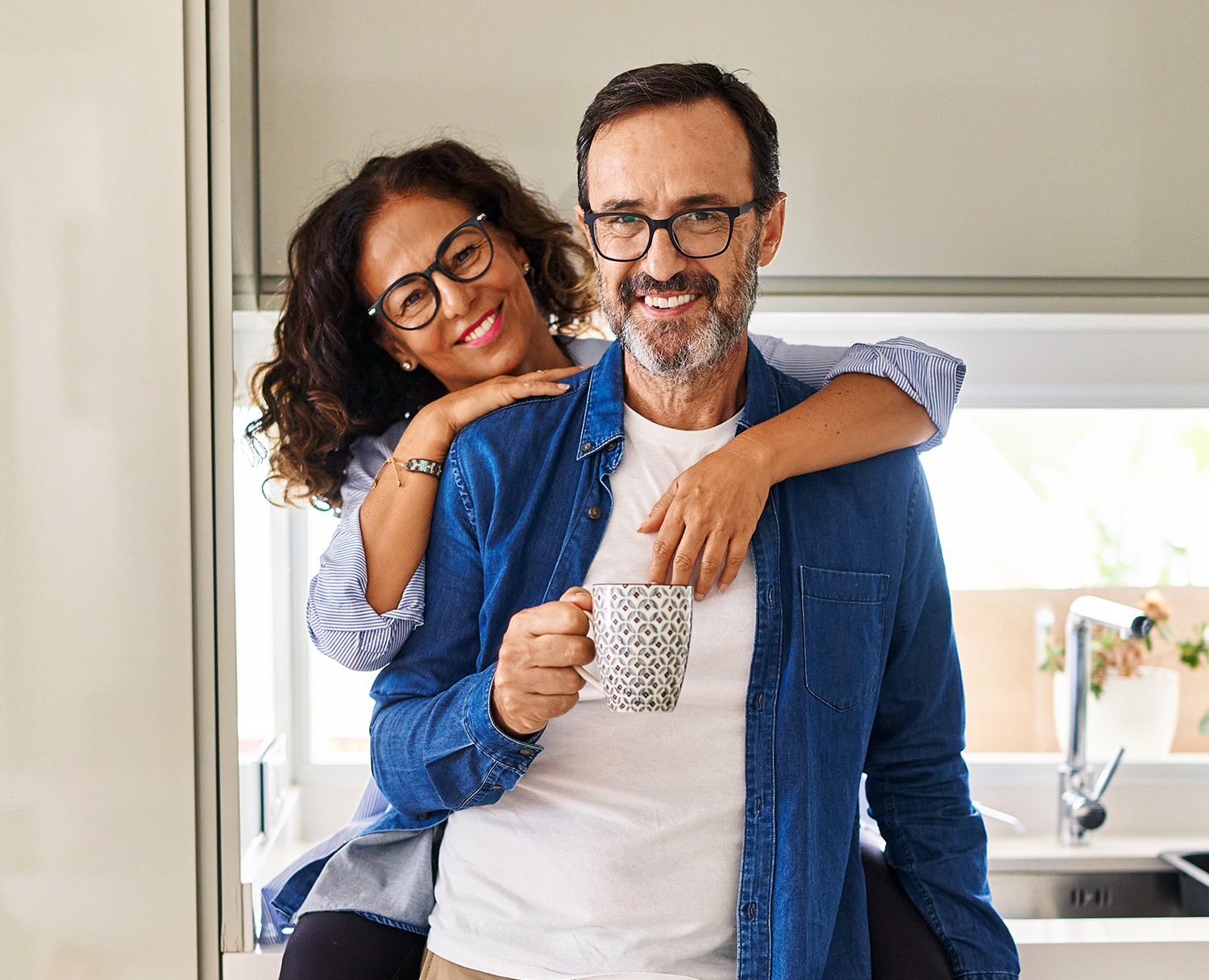 A smiling couple standing in the kitchen, with the woman playfully embracing the man from behind as he holds a coffee mug. Both are wearing glasses and casual clothing - Eye Doctor Rolla MO | Invision Family Eye Care