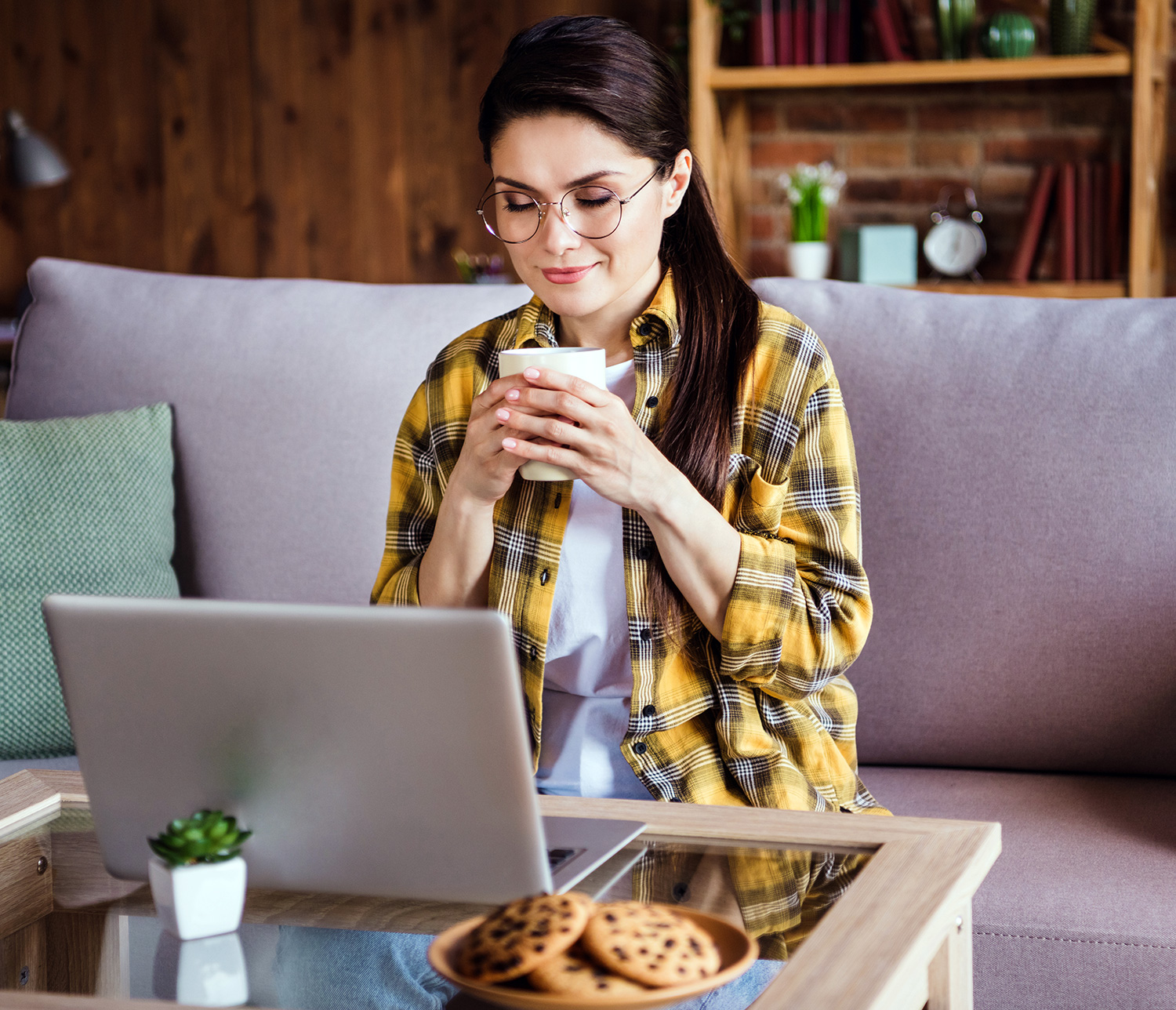A woman wearing glasses and a yellow plaid shirt sits on a couch, holding a cup while looking at her laptop. A plate of cookies is on the table in front of her - Eye Doctor Rolla MO | Invision Family Eye Care