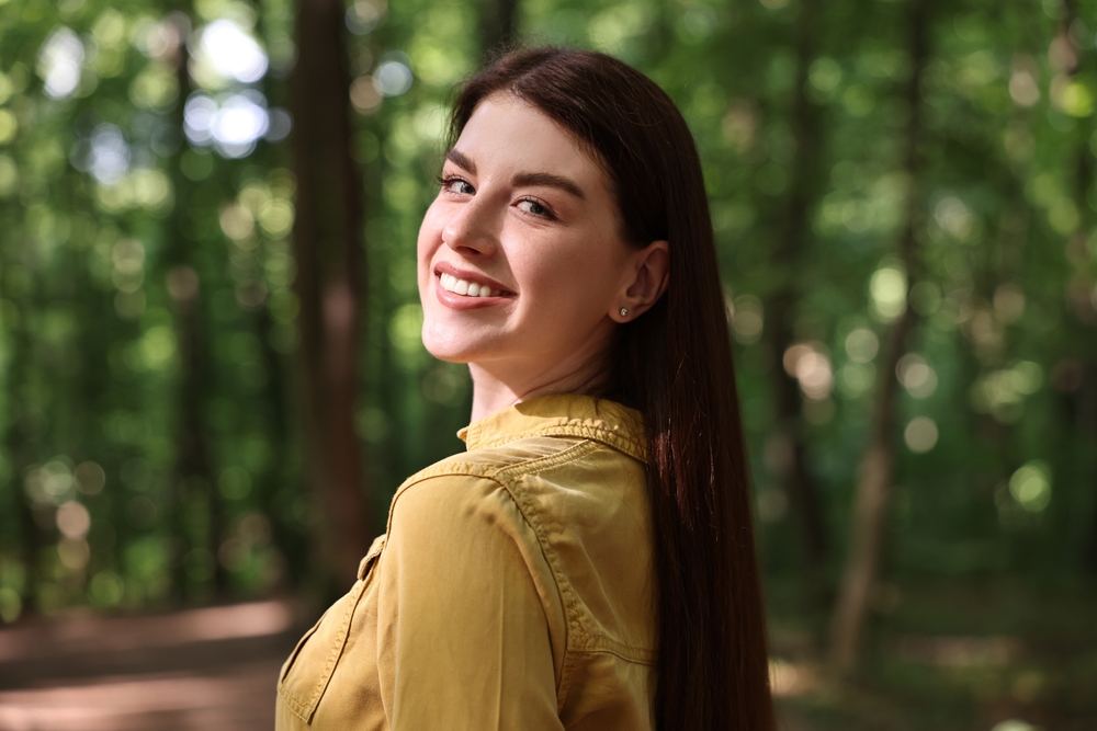 A woman with a bright, natural smile turns toward the camera while walking through a lush forest. The soft, green background enhances the serene, healthy, and vibrant look, representing clarity and wellness – Cataract Treatment.